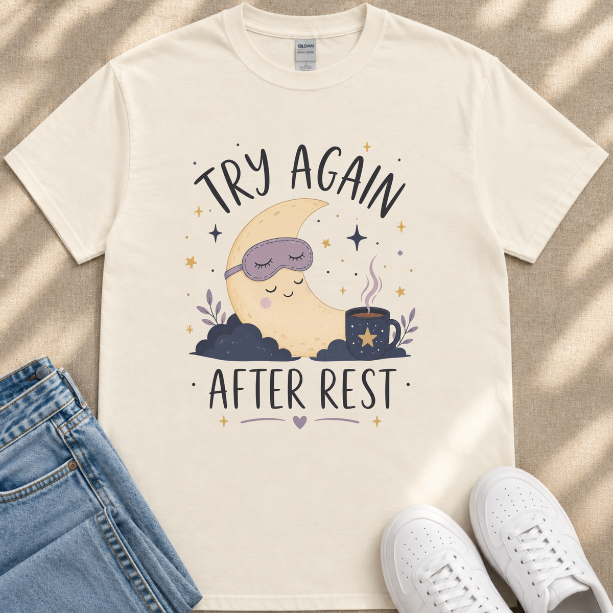 A white t-shirt with a graphic design depicting a sleeping crescent moon and stars, along with the text "TRY AGAIN AFTER REST" and a mug with a star.