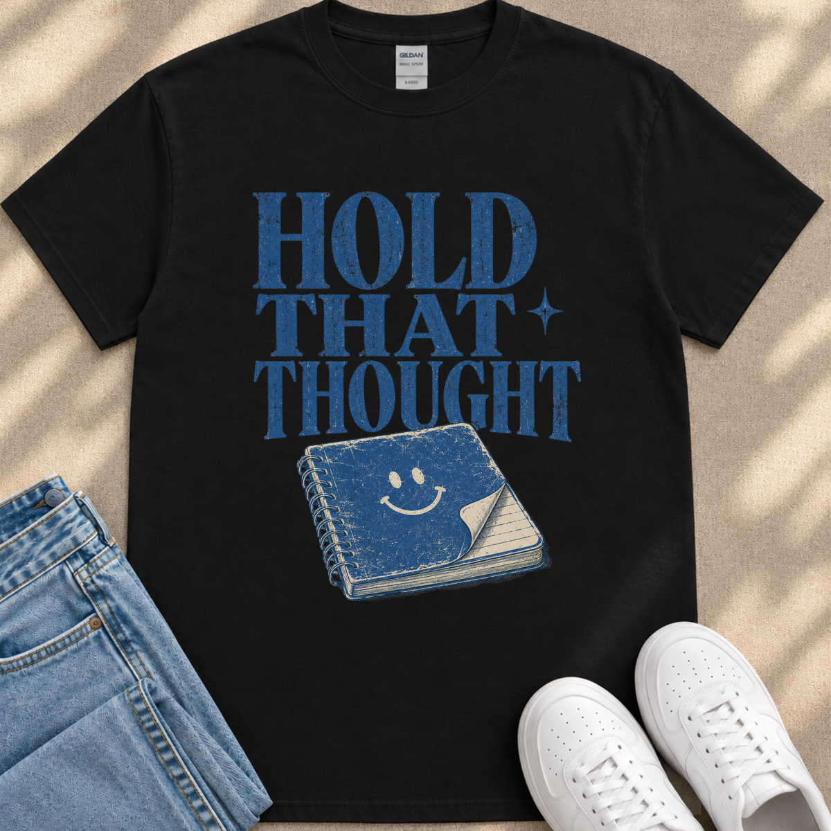 A black t-shirt with a graphic design featuring a blue book with a smiley face on the cover, along with the text "HOLD THAT THOUGHT" printed on it. The t-shirt is placed on a beige surface, and there are a pair of blue jeans and white sneakers visible in the image.