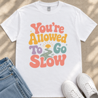 A white t-shirt with a colorful graphic design that says "You're Allowed to Go Slow" printed on it.