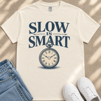 A white t-shirt with the text "SLOW IS SMART" printed on it, along with a clock graphic. The t-shirt is placed on a beige surface, and there are a pair of jeans and white sneakers visible in the image.