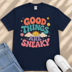 A navy blue t-shirt with a colorful graphic design that says "GOOD THINGS ARE SNAKY" and features various cheerful elements such as clouds, stars, and smiley faces.
