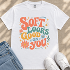 A white t-shirt with a colorful graphic design that says "Soft looks good on you" surrounded by flowers and stars.