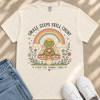 A white t-shirt with a colorful graphic design depicting a frog sitting on a mushroom, surrounded by flowers and a rainbow. The text on the t-shirt reads "Small steps still count" and "Even the mobbly ones".