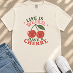 a white t-shirt with a graphic design of two cherries and the text "Life is weird. Have a cherry." The t-shirt is placed on a beige surface, and there are a pair of blue jeans and white sneakers visible in the background.