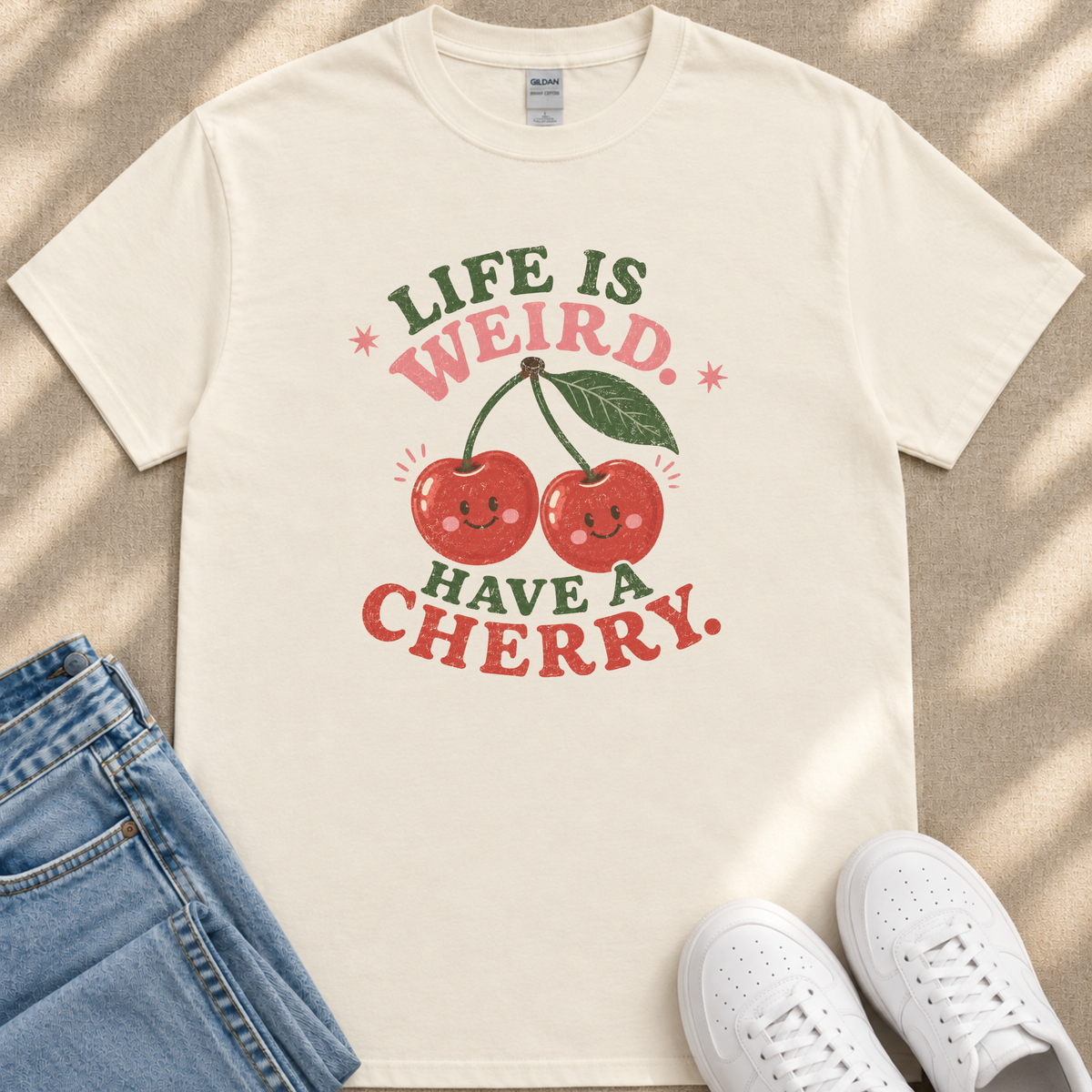 a white t-shirt with a graphic design of two cherries and the text "Life is weird. Have a cherry." The t-shirt is placed on a beige surface, and there are a pair of blue jeans and white sneakers visible in the background.