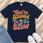 A navy blue t-shirt with a colorful graphic design that says "You're Allowed to Go Slow" in a retro style.