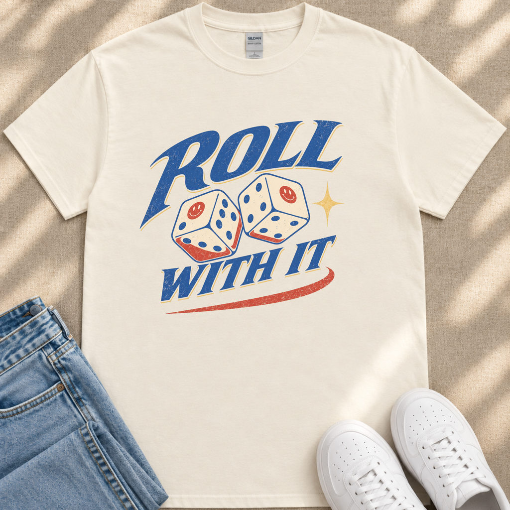 A white t-shirt with a graphic design featuring two dice with smiley faces, along with the text "ROLL WITH IT" and a red and blue stripe. The t-shirt is placed on a beige surface, and there are a pair of blue jeans and white sneakers visible in the image.