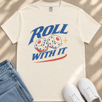 A white t-shirt with a graphic design featuring two dice with smiley faces, along with the text "ROLL WITH IT" and a red and blue stripe. The t-shirt is placed on a beige surface, and there are a pair of blue jeans and white sneakers visible in the image.