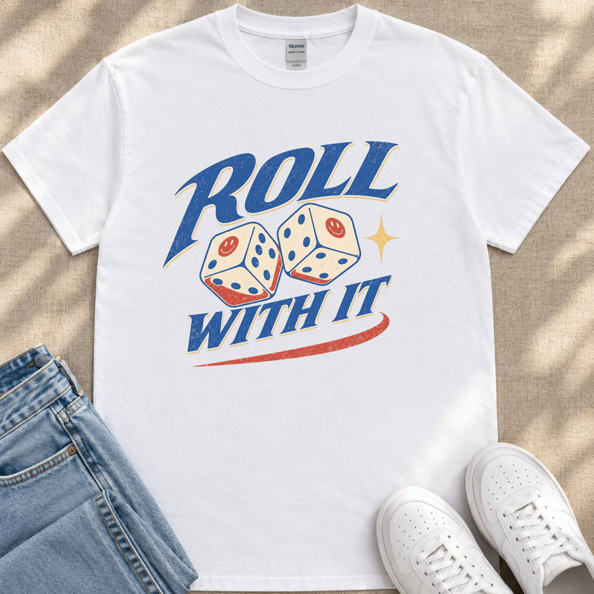 A white t-shirt with a graphic design featuring two dice with smiley faces, along with the text "ROLL WITH IT" and a red and yellow star. The t-shirt is placed on a beige surface, and there are a pair of blue jeans and white sneakers visible in the image.