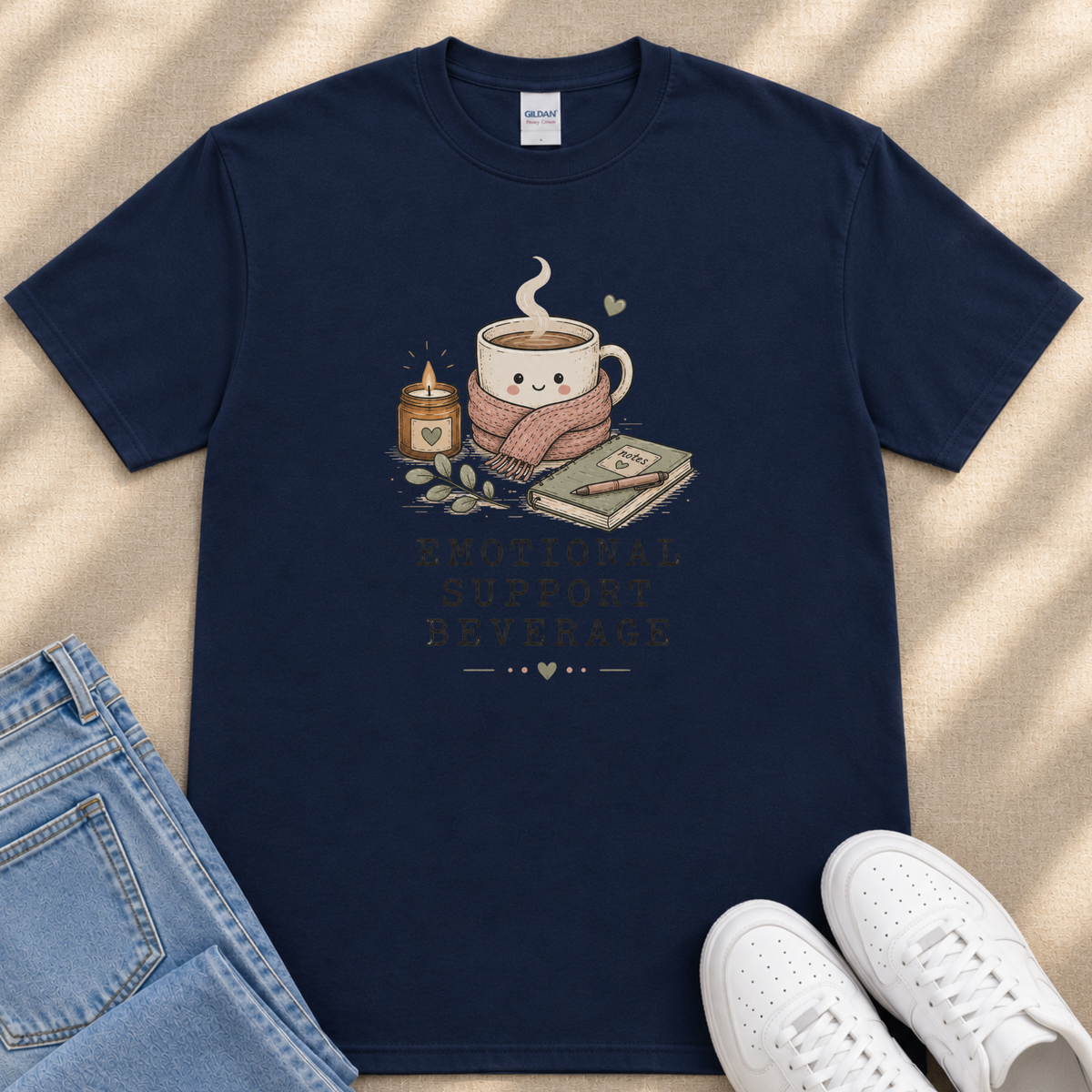 A navy blue t-shirt with a graphic design depicting a cup of coffee, a candle, and a book, along with the text "Emotional Support Beverage" printed on it.