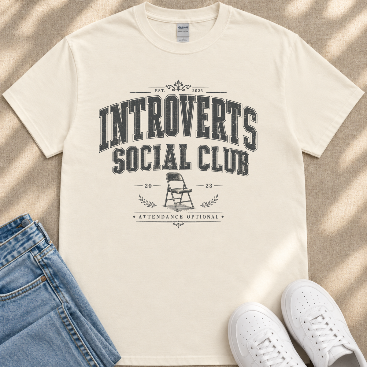 A beige t-shirt with a graphic design and text, along with a pair of jeans and white sneakers, laid out on a surface.