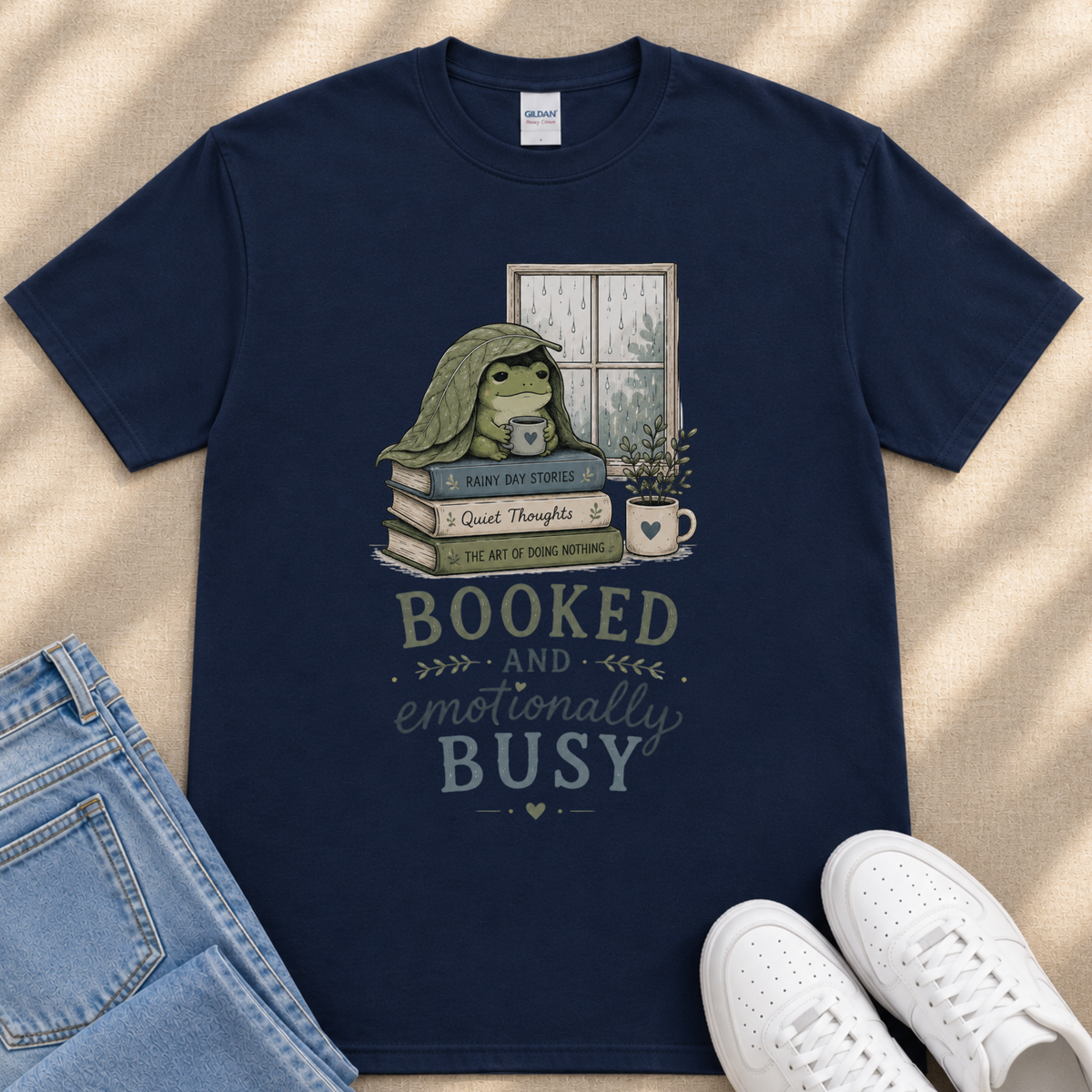 A navy blue t-shirt with a graphic design depicting a frog sitting on a stack of books, with the text "BOOKED AND emotionally BUSY" printed on it.