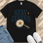 a black t-shirt with a graphic design of a magnifying glass and a smiling star, along with the text "STAY CURIOUS" printed on it. The t-shirt is placed on a beige surface, and there are a pair of blue jeans and white sneakers visible in the image.