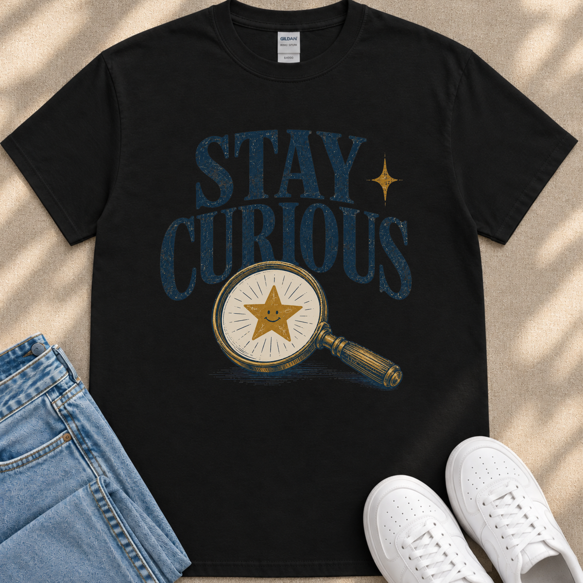 a black t-shirt with a graphic design of a magnifying glass and a smiling star, along with the text "STAY CURIOUS" printed on it. The t-shirt is placed on a beige surface, and there are a pair of blue jeans and white sneakers visible in the image.