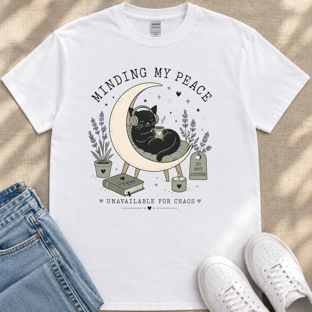A white t-shirt with a graphic design depicting a black cat sitting on a crescent moon, surrounded by potted plants and flowers, with the text "MINDING MY PEACE" and "UNAVAILABLE FOR CHAOS" printed on it.