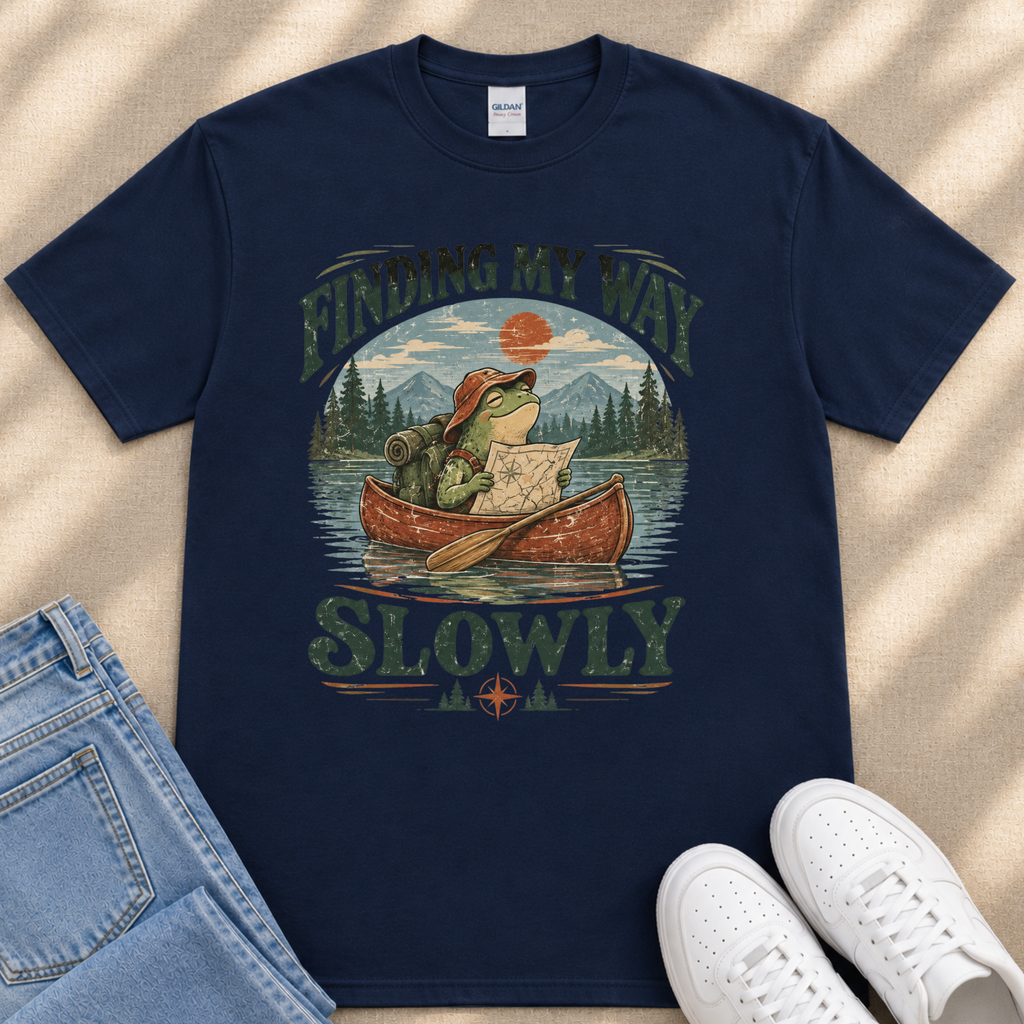 A navy blue t-shirt with a graphic design depicting a frog in a canoe on a lake, with the text "FINDING MY WAY SLOWLY" printed on it.