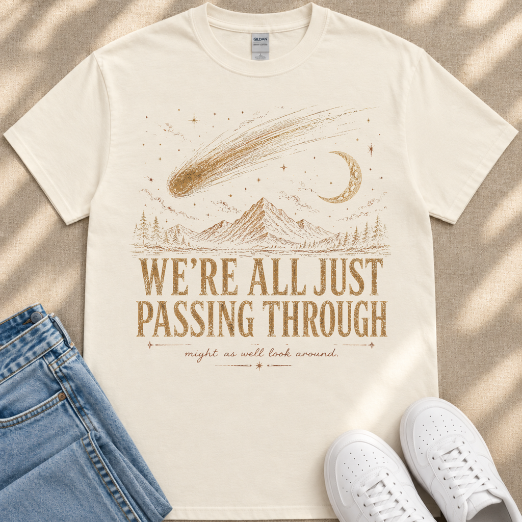 A white t-shirt with a graphic design depicting a shooting star, mountains, and a crescent moon, along with the text "WE'RE ALL JUST PASSING THROUGH" and "mighty as well look around." The t-shirt is laid out on a beige surface, and there are a pair of blue jeans and white sneakers visible in the image.
