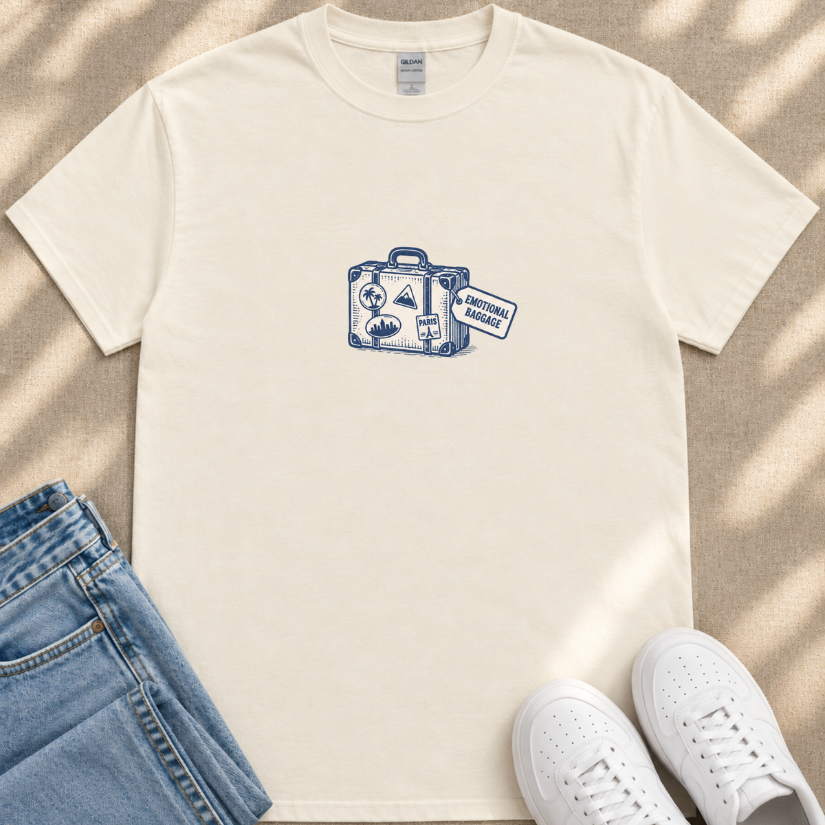 A white t-shirt with a graphic of a suitcase and a blue and white label that says "EMOTIONAL BAGGAGE" is displayed on a beige surface, along with a pair of blue jeans and white sneakers.