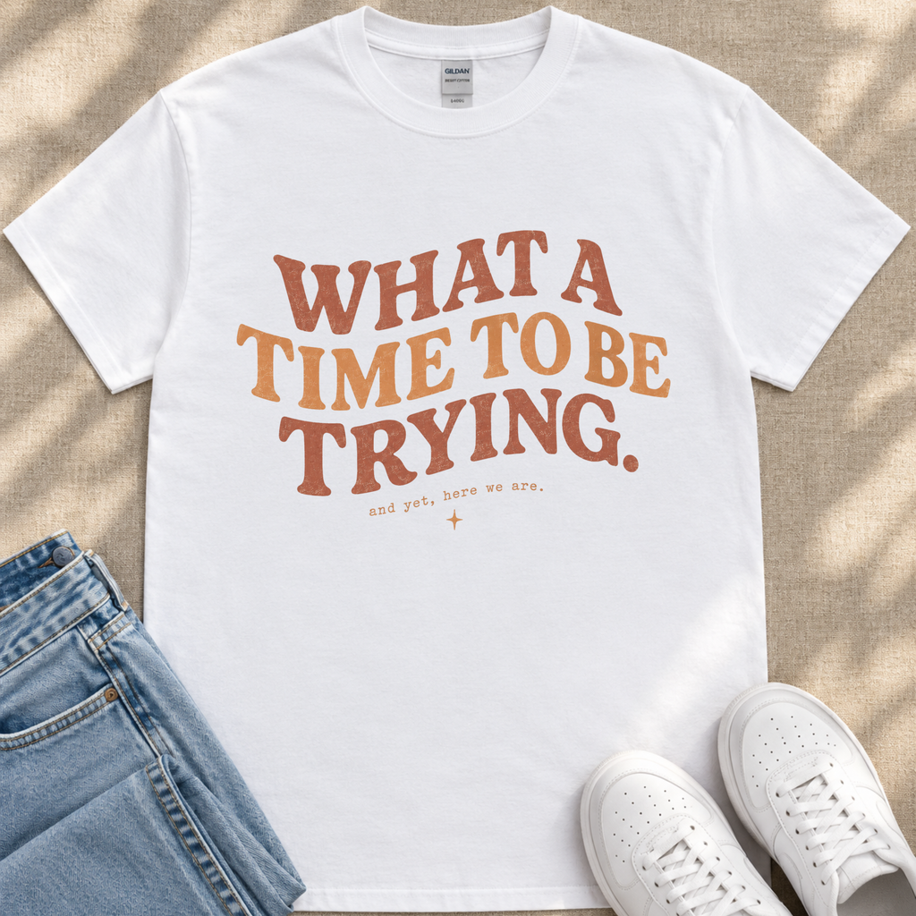 A white t-shirt with the text "WHAT A TIME TO BE TRYING" printed on it, along with the phrase "and get, here we are." placed on a pair of jeans and white sneakers.