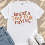 A white t-shirt with the text "WHAT A TIME TO BE TRYING" printed on it, along with the phrase "and get, here we are." placed on a pair of jeans and white sneakers.