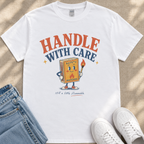 A white t-shirt with a graphic design featuring a cartoon matchbox character holding a lighter and the text "HANDLE WITH CARE" and "SAFETY MATCHES" printed on it.