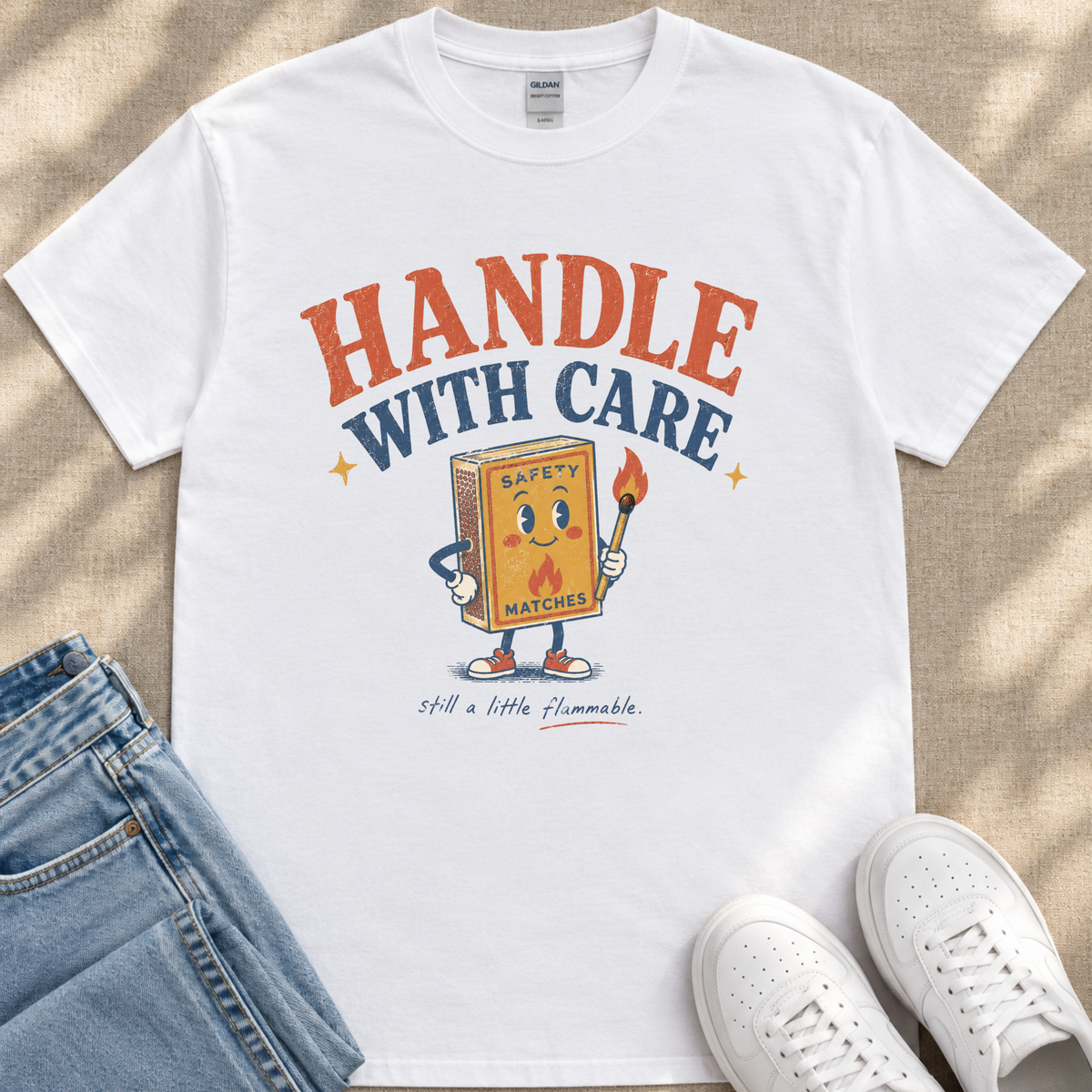 A white t-shirt with a graphic design featuring a cartoon matchbox character holding a lighter and the text "HANDLE WITH CARE" and "SAFETY MATCHES" printed on it.