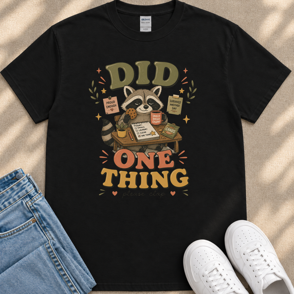 a black t-shirt with a colorful graphic design depicting a raccoon sitting at a desk with various items, such as a book, a cup, and a plant. The text on the t-shirt reads "Did One Thing" and includes a quote about being productive. The t-shirt is placed on a beige surface, and there are a pair of jeans and white sneakers visible in the background.