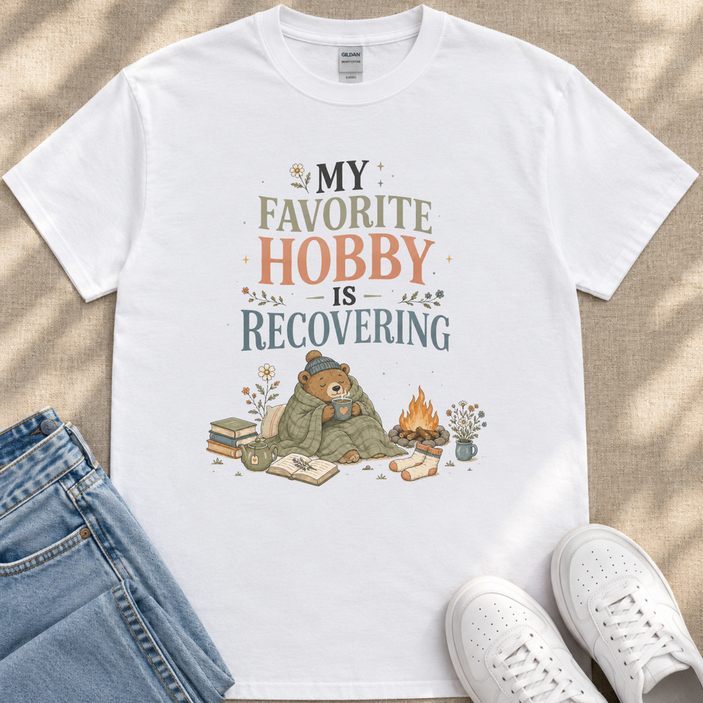 A white t-shirt with a graphic design depicting a bear wrapped in a blanket, sitting next to a campfire and books, along with the text "My favorite hobby is recovering".