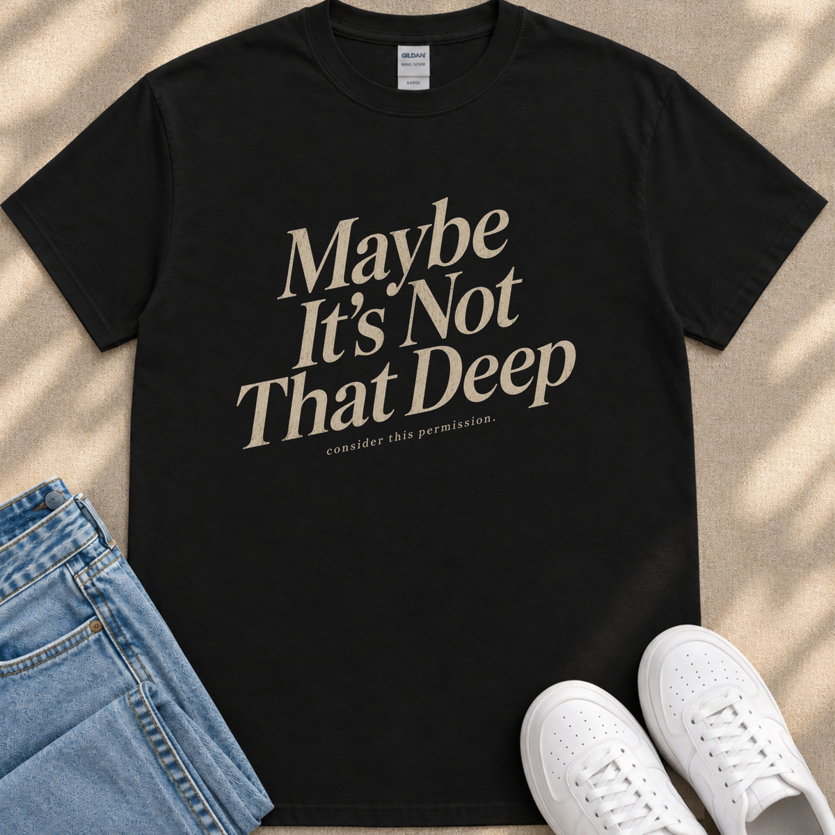 A black t-shirt with a printed message, "Maybe It's Not That Deep", along with the text "Considering this permission" printed below it. The t-shirt is placed on a beige surface, and there are a pair of blue jeans and white sneakers visible in the image.