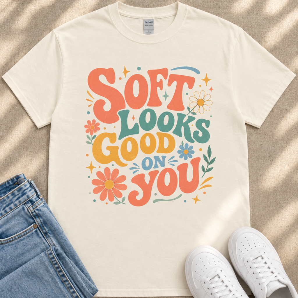 A white t-shirt with a colorful graphic design that says "Soft looks good on you" surrounded by flowers and stars. The t-shirt is placed on a beige surface, and there are a pair of blue jeans and white sneakers visible in the image.