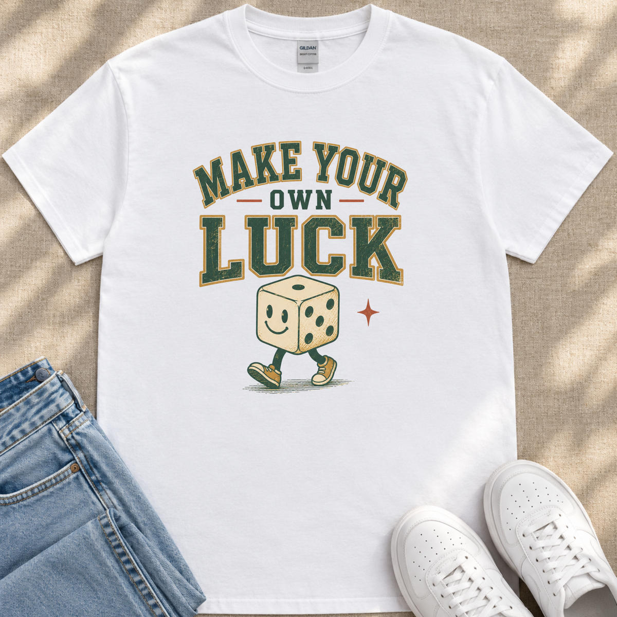 A white t-shirt with a graphic design featuring a smiling dice character and the text "MAKE YOUR OWN LUCK" printed on it.