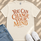 A white t-shirt with the text "You Can Change Your Mind" printed on it, along with the phrase "that's allowed." The t-shirt is laid out on a beige surface, and there are a pair of blue jeans and white sneakers visible in the image.