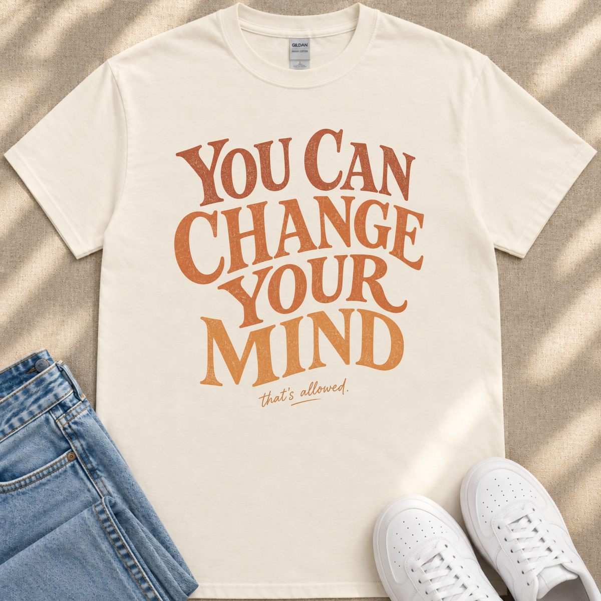 A white t-shirt with the text "You Can Change Your Mind" printed on it, along with the phrase "that's allowed." The t-shirt is laid out on a beige surface, and there are a pair of blue jeans and white sneakers visible in the image.
