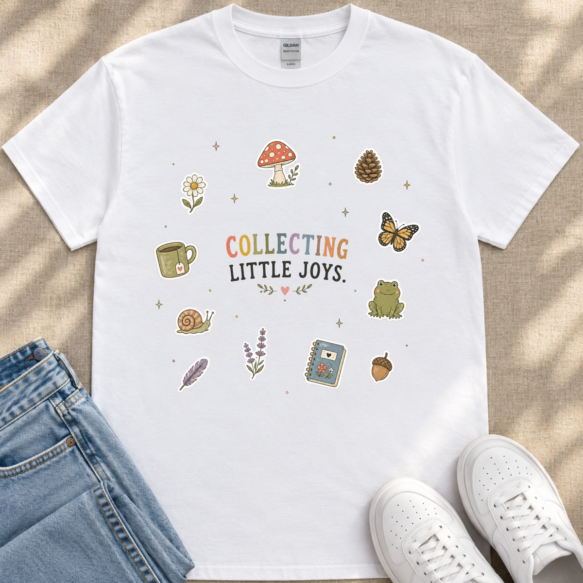 A white t-shirt with a colorful graphic design featuring various objects such as a mushroom, flower, butterfly, cup, and more, along with the text "COLLECTING LITTLE JOYS." The t-shirt is placed on a beige surface, and there are a pair of jeans and white sneakers visible in the image.