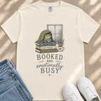 A white t-shirt with a graphic design depicting a frog sitting on a stack of books, with the text "BOOKED AND emotionally BUSY" below the image.