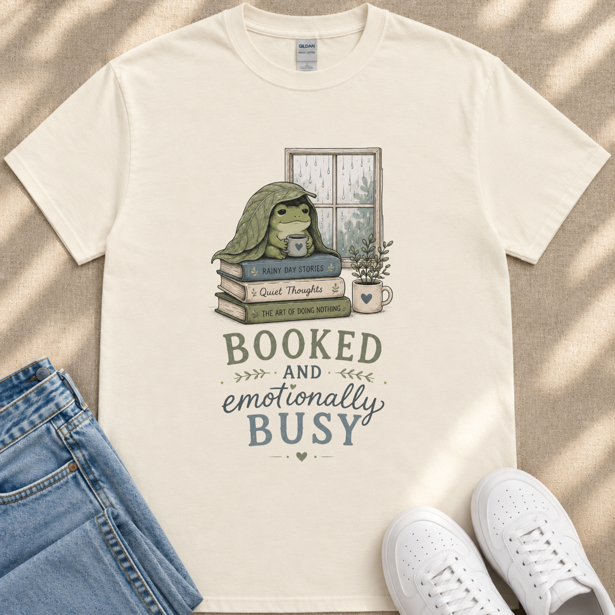 A white t-shirt with a graphic design depicting a frog sitting on a stack of books, with the text "BOOKED AND emotionally BUSY" below the image.