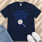 A navy blue t-shirt with the text "FIND SOMETHING GOOD" and an illustration of a magnifying glass with a star on it, along with the text "It's usually around somewhere." The t-shirt is laid out on a beige surface, and there are a pair of jeans and white sneakers visible in the image.