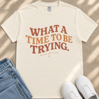 A white t-shirt with the text "What a TIME to BE TRYING." printed on it, along with the phrase "and get, here we are." placed on a beige surface.