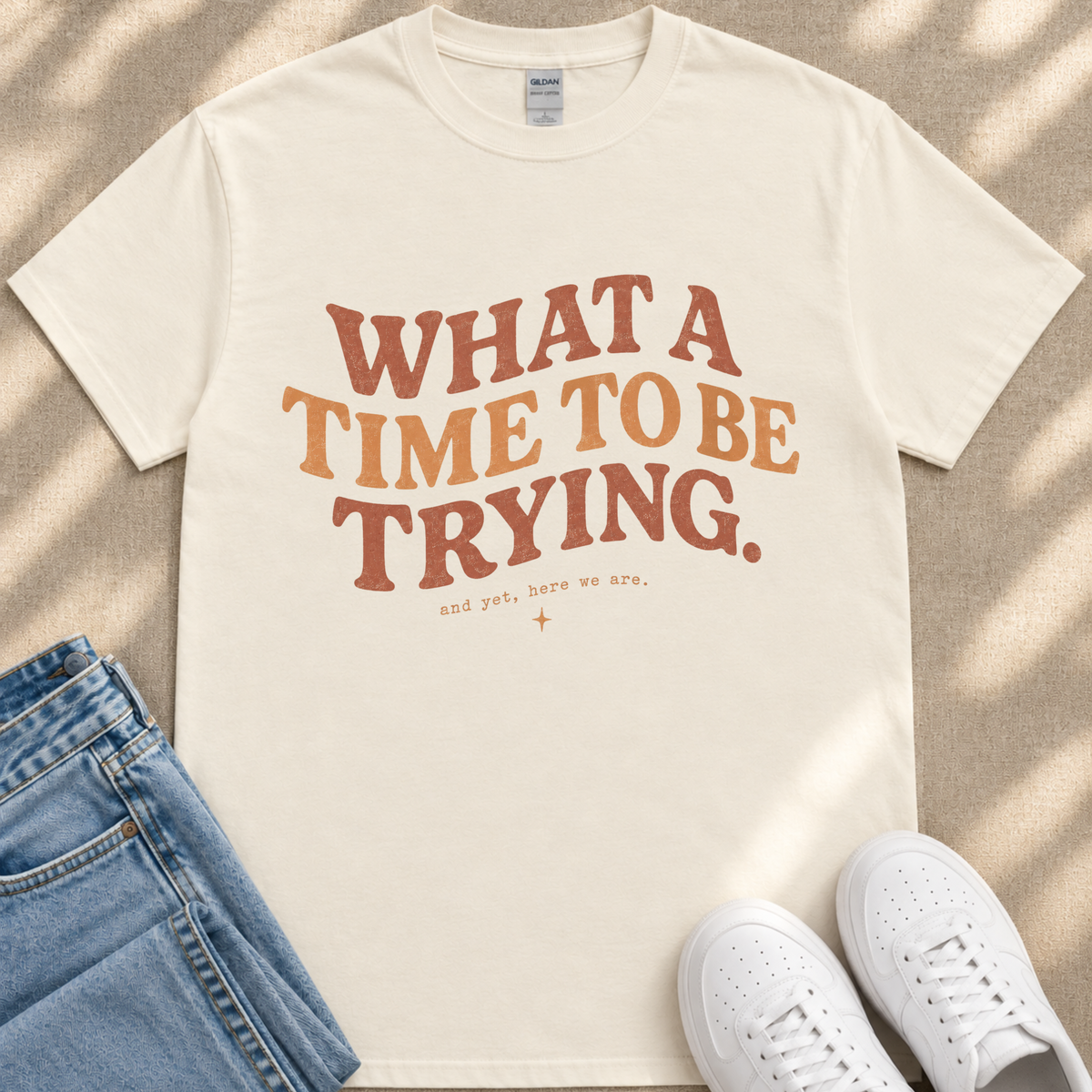 A white t-shirt with the text "What a TIME to BE TRYING." printed on it, along with the phrase "and get, here we are." placed on a beige surface.