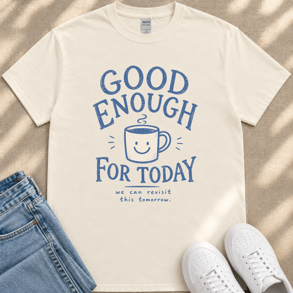 A white t-shirt with a blue graphic design that says "GOOD ENOUGH FOR TODAY" and a smiling coffee mug. The t-shirt is placed on a beige surface, and there are a pair of blue jeans and white sneakers visible in the image.
