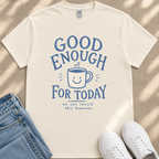 A white t-shirt with a blue graphic design that says "GOOD ENOUGH FOR TODAY" and a smiling coffee mug. The t-shirt is placed on a beige surface, and there are a pair of blue jeans and white sneakers visible in the image.