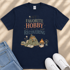 A navy blue t-shirt with a graphic design depicting a bear wrapped in a blanket, sitting by a campfire and surrounded by books and flowers. The text on the t-shirt reads "My Favorite Hobby is Recovering".