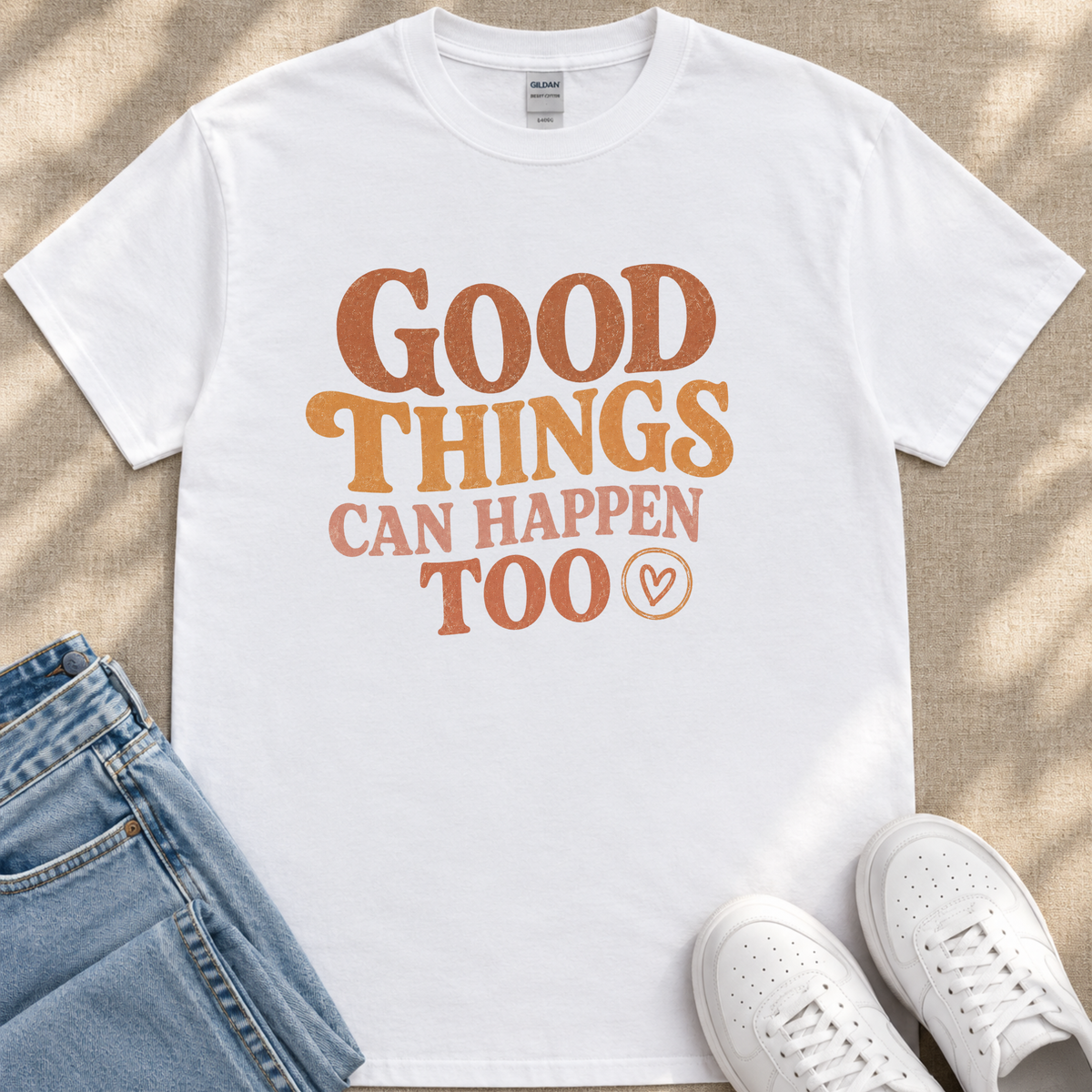 A white t-shirt with the text "GOOD THINGS CAN HAPPEN TO" printed on it, along with a heart symbol. The t-shirt is placed on a beige surface, and there are a pair of jeans and white sneakers visible in the image.