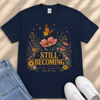 A navy blue t-shirt with a floral design and the text "STILL BECOMING" printed on it, along with a pair of jeans and white sneakers.