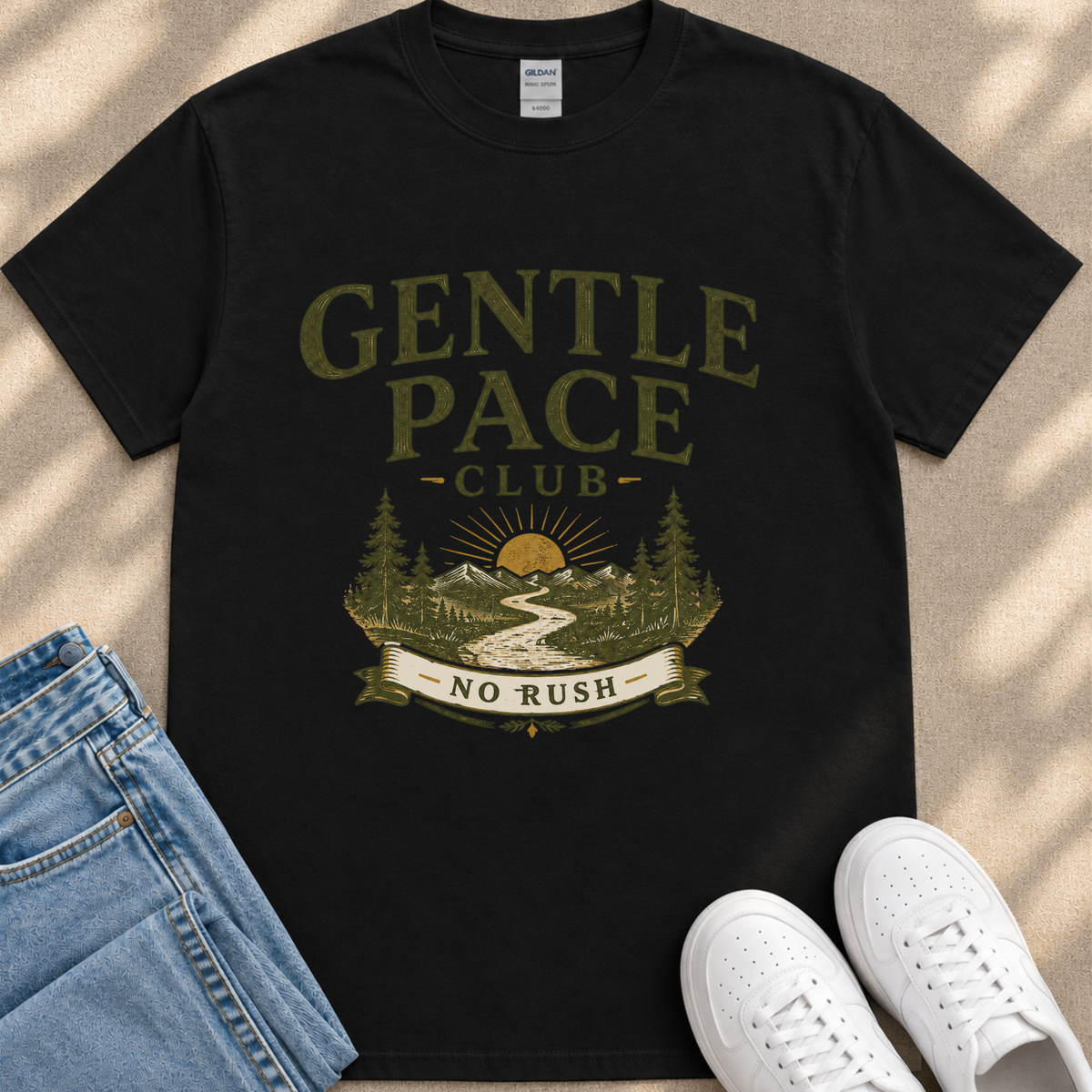 A black t-shirt with a graphic design featuring a mountain landscape, a sun, and a banner that says "GENTLE PACE CLUB NO RUSH". The t-shirt is placed on a beige surface, and there are a pair of blue jeans and white sneakers visible in the image.