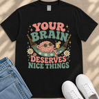 A black t-shirt with a colorful graphic design featuring a cartoon brain character in a hammock, surrounded by flowers and text that reads "Your brain deserves nice things".