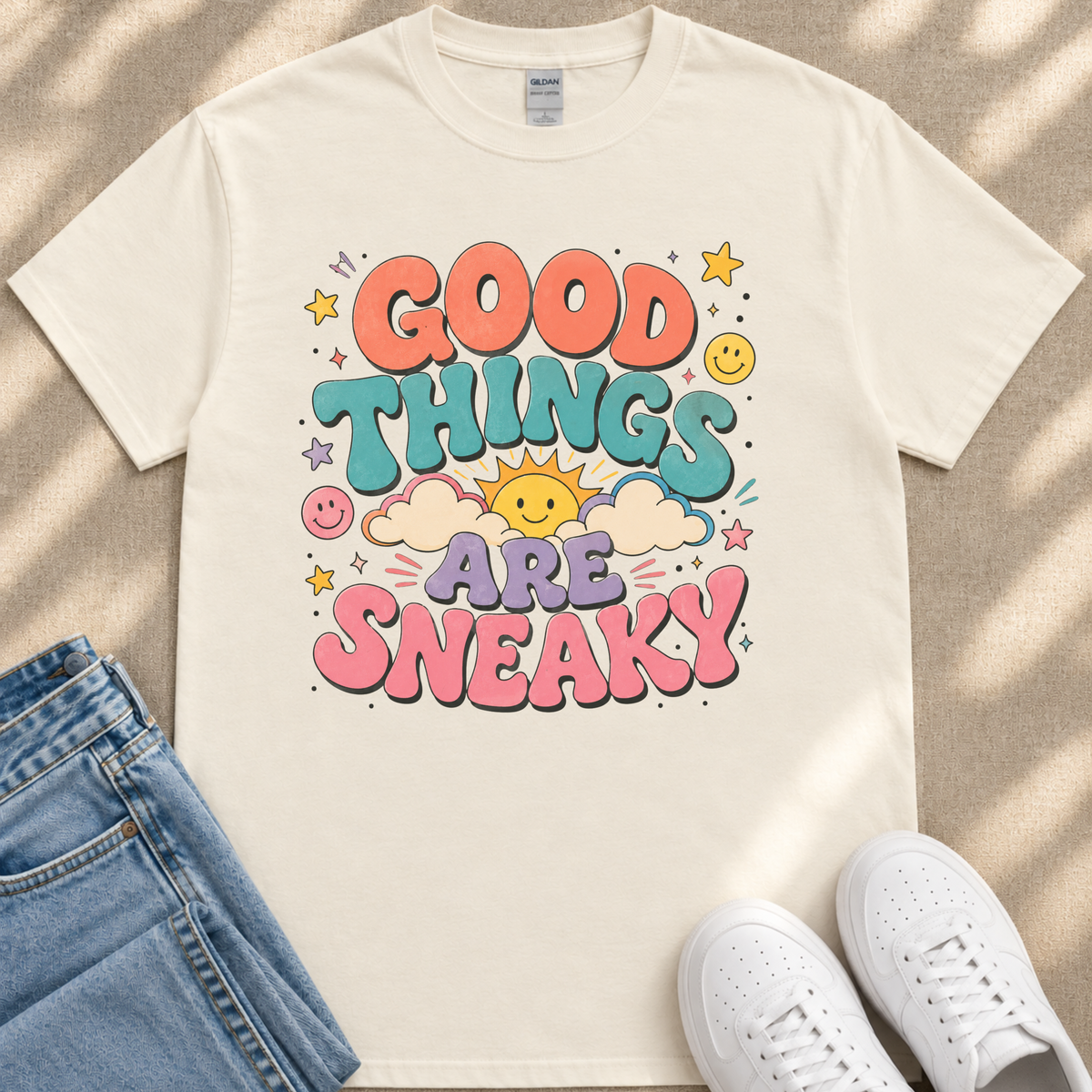 A white t-shirt with a colorful graphic design that says "GOOD THINGS ARE SNAKY" and includes various cheerful elements such as smiley faces, clouds, and stars.