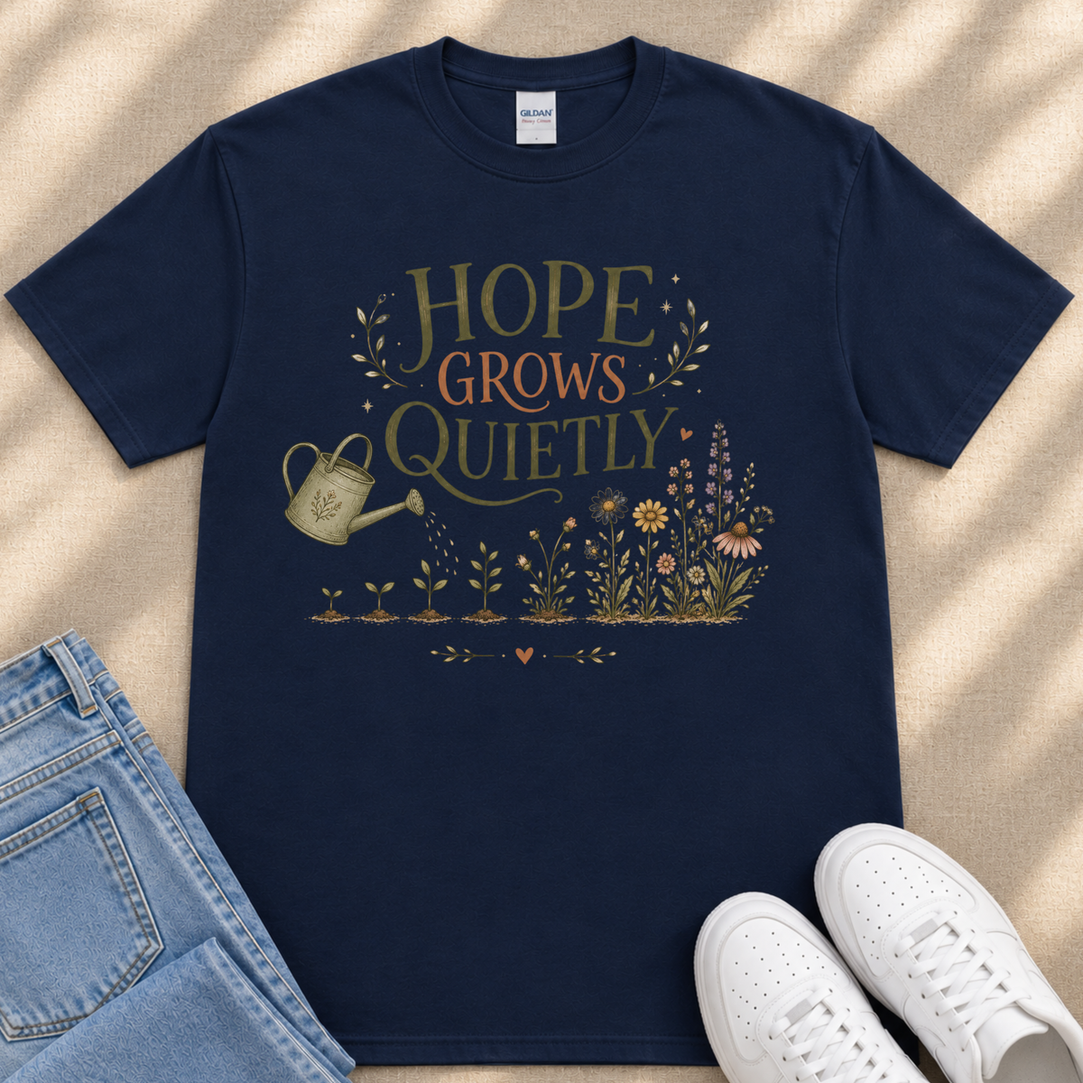 A navy blue t-shirt with a printed design of a watering can surrounded by flowers and plants, along with the text "HOPE GROWS QUICELY". The t-shirt is placed on a beige surface, and there are a pair of jeans and white sneakers visible in the image.