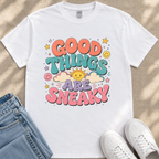 A white t-shirt with a colorful graphic design that says "GOOD THINGS ARE SNAKY" surrounded by various cheerful elements such as smiley faces, clouds, and stars.