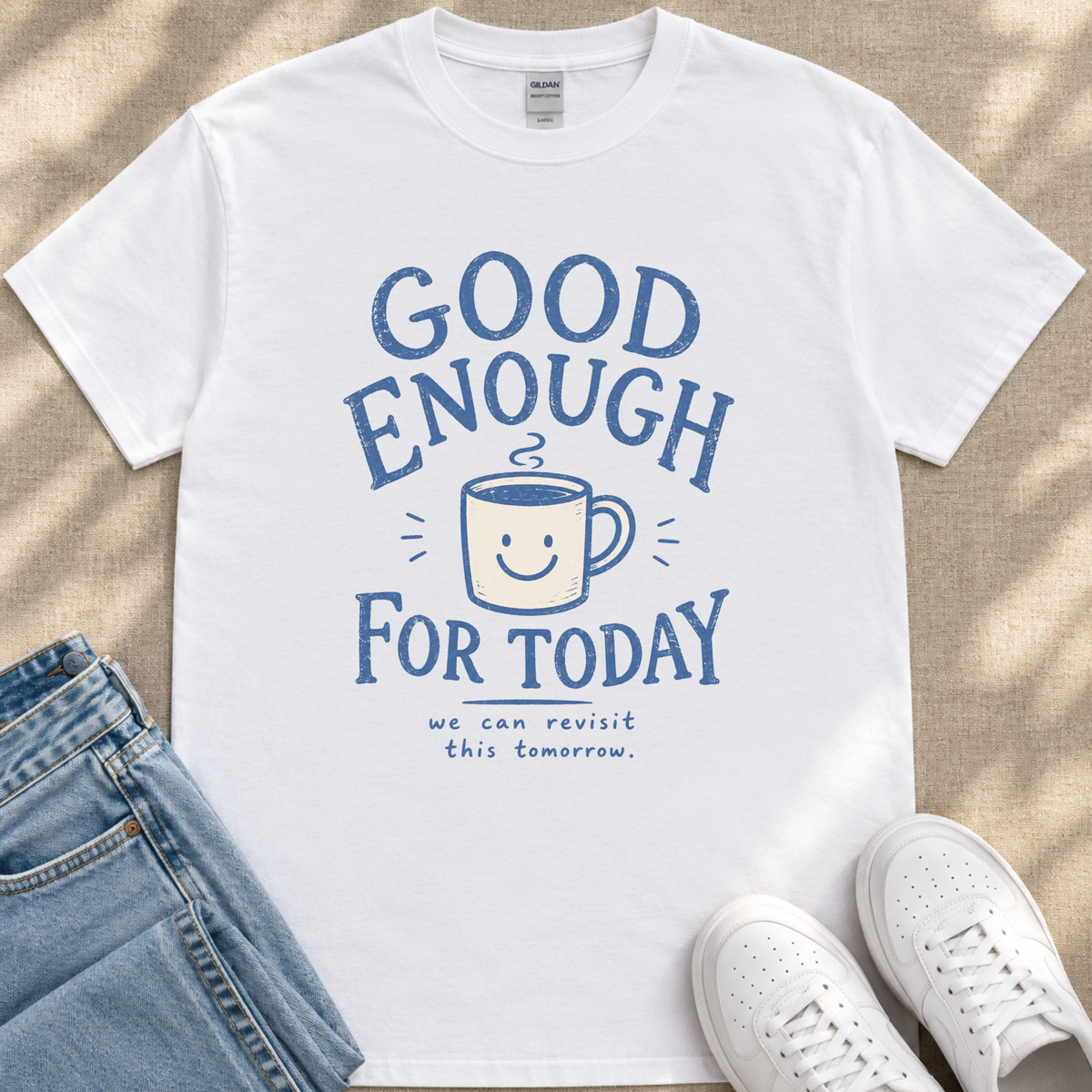A white t-shirt with a blue graphic design that says "GOOD ENOUGH FOR TODAY" and a coffee cup with a smiley face. Below the graphic, there is a smaller text that says "we can revisit this tomorrow."